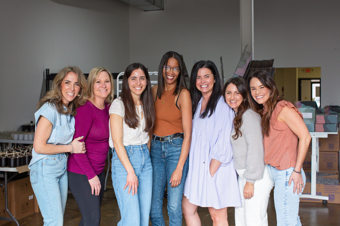 Seven women stand close together, posing in a warehouse with laurelboxes and gifts in the background.