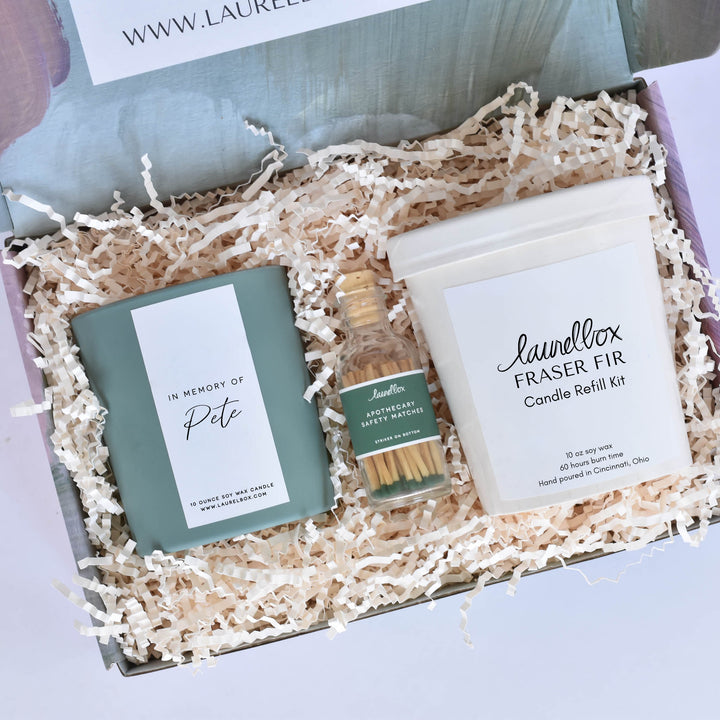 A close-up of a laurelbox gift set with a green candle with a white label with the text "In Memory of Pete", a glass container with a green label featuring the Apothecary Safety Matches text, filled with green matches, and a laurelbox Fraser Fir candle refill kit.
