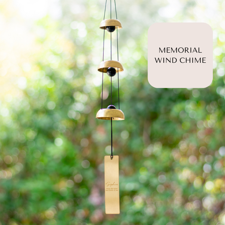 A wind chime hanging outside that features three metallic bells, black cords, and a vertical rectangular tag at the bottom with the text Sophia and Hear the wind and think of me