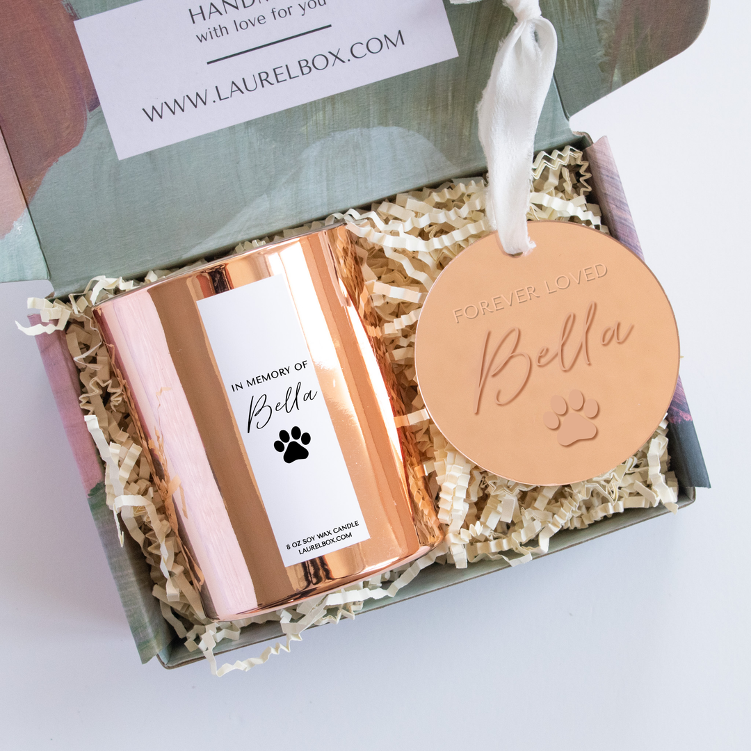 A close-up of a laurelbox gift set with a gold candle with a white label with the text "In Memory of Bella" with a paw print, and a circular gold ornament with the text "Forever Loved Bella" with a paw print engraved with a white silk ribbon at the top.
