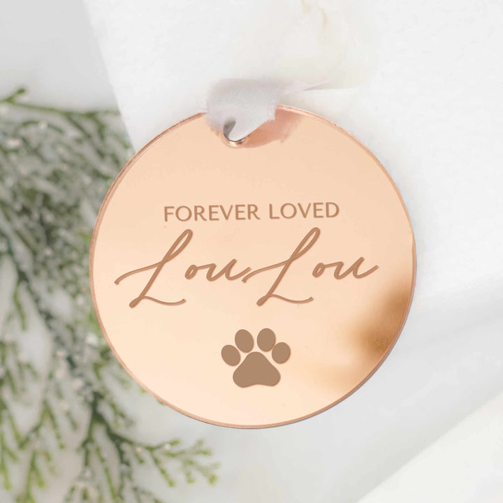 A personalized gold mirrored ornament featuring the words "Forever Loved" and "Lou Lou" with a paw print symbol engraved and a white ribbon at the top is set against a white background with pine branches on the left.