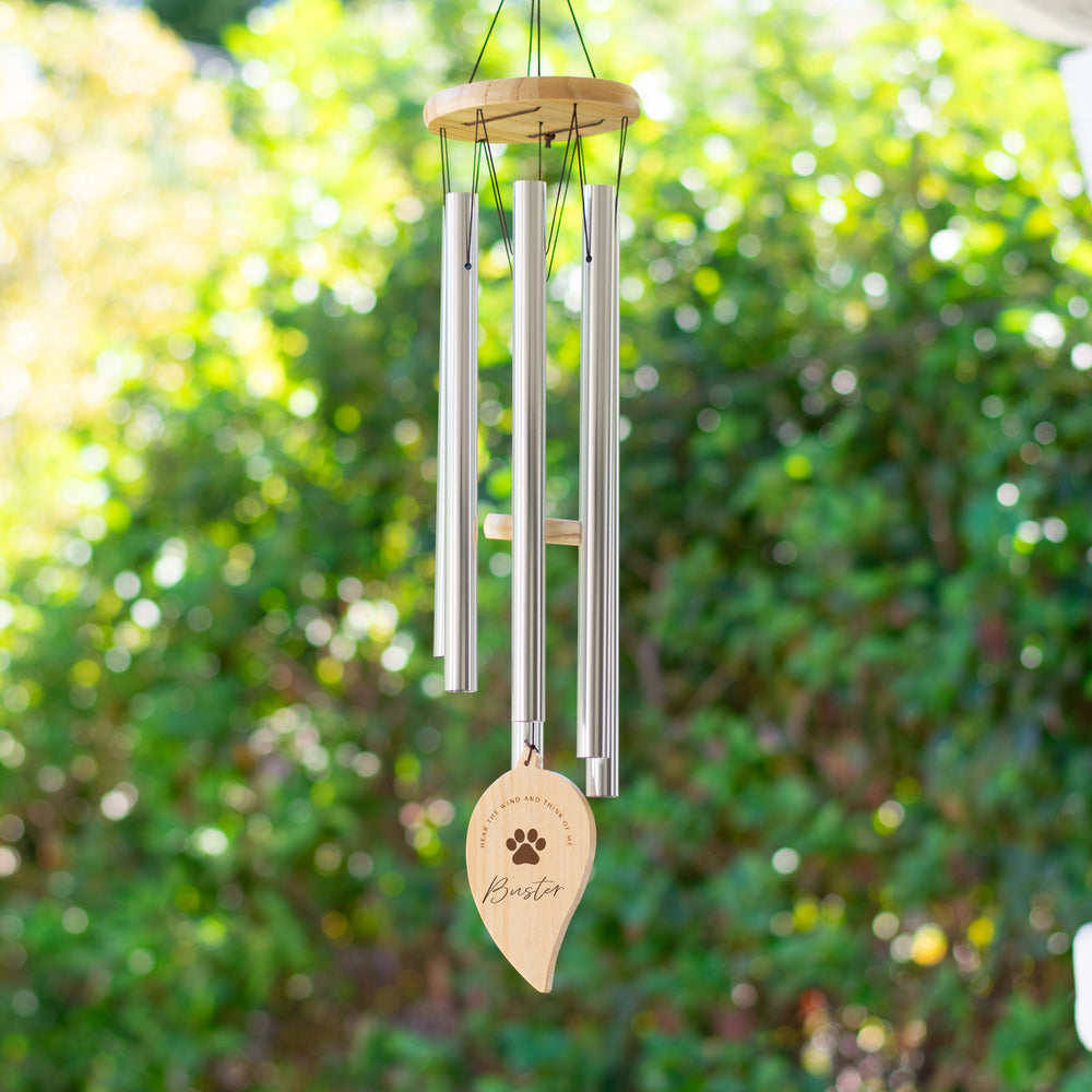 A wind chime handing outdoors features long metal tubes with a wooden disc at the top. A wooden leaf is at the bottom with a paw print and the text Buster and Hear the wind and think of me
