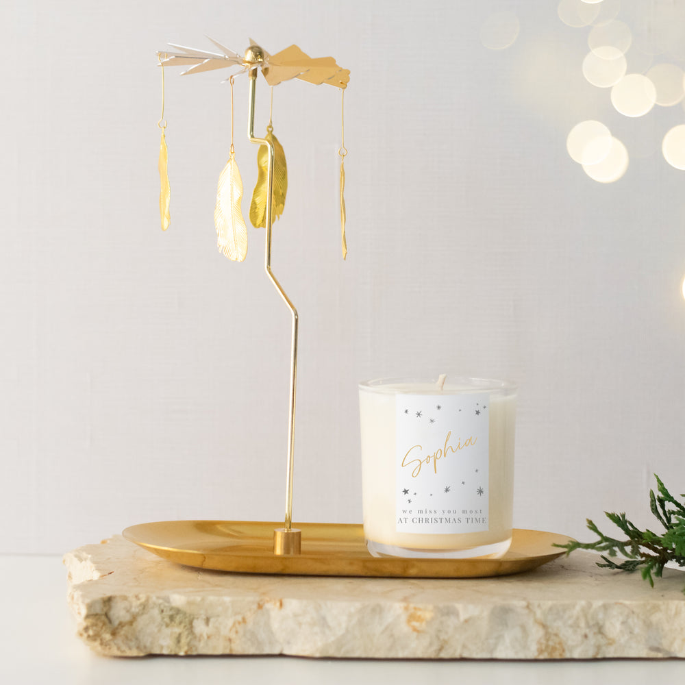 A gold metal rotary stand featuring four feather pendants and a gold tray placed on a marble surface. A customized candle is sitting on the gold rotary stand tray.