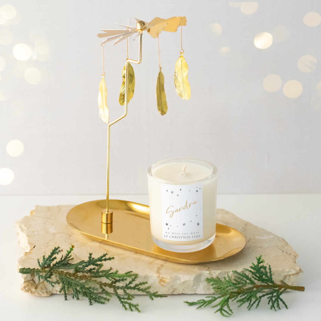 An angled view of a gold metal rotary stand featuring four feather pendants and a gold tray placed on a marble surface. A customized candle is sitting on the gold rotary stand tray.