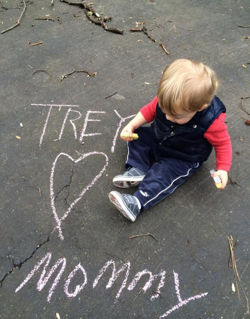 photo of a small blonde child sitting on pavement holding chalk sticks with the words "Trey Hearts Mommy" written in pink chalk next to him