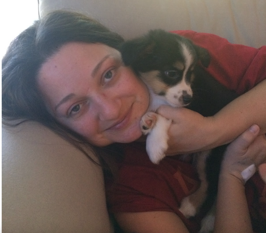 photo of a woman with brown hair and blue eyes wearing a red shirt, laying down on a tan couch holding a small multi colored puppy