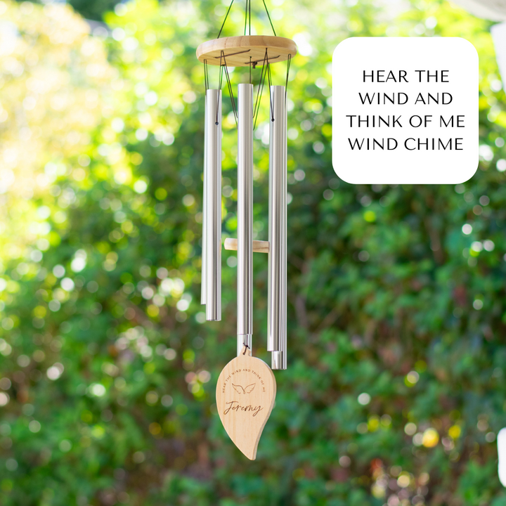 A wind chime handing outdoors features long metal tubes with a wooden disc at the top. A wooden leaf is at the bottom with the text Jeremy and Hear the wind and think of me