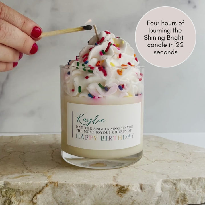 A woman lighting a glass candle labeled Kaylee May the angels sing to you the most joyous chorus of Happy Birthday with whipped cream and sparkles at the top. Sitting on a granite surface.