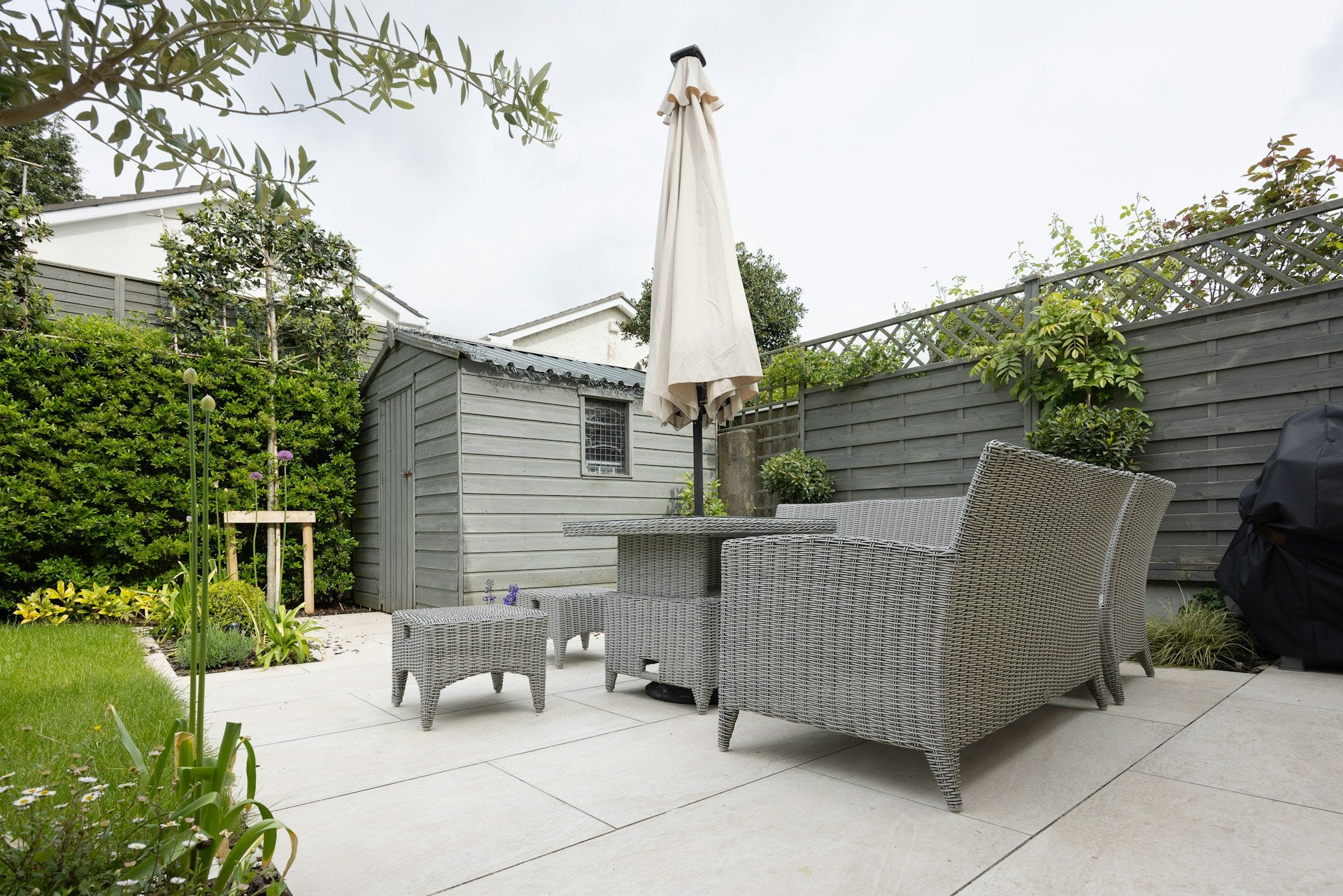 photo of a backyard with grey wicker table, chair and couch on top of stones surrounded by a fence, shed and a green garden