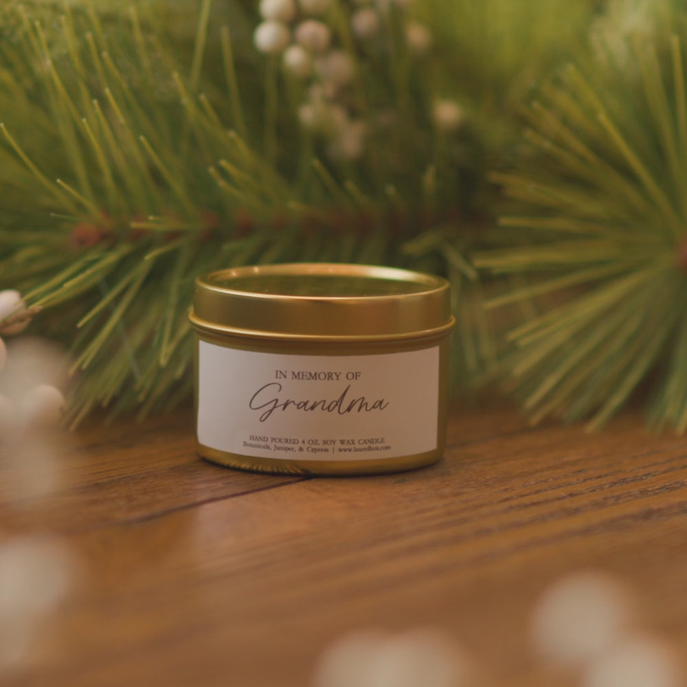 A woman holding up a gold-coated wide candle with a white sticker with the text in Memory of Grandma sitting on a wooden table
