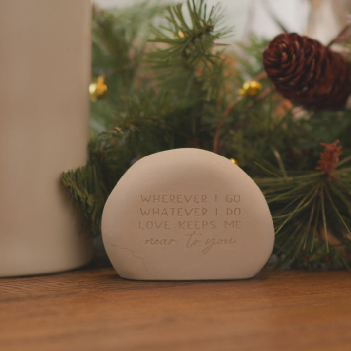 A memorial gemstone with Whenever I Go Whatever I Do Love Keeps Me Near To You is engraved on it, sitting next to pine needles and pine cones.