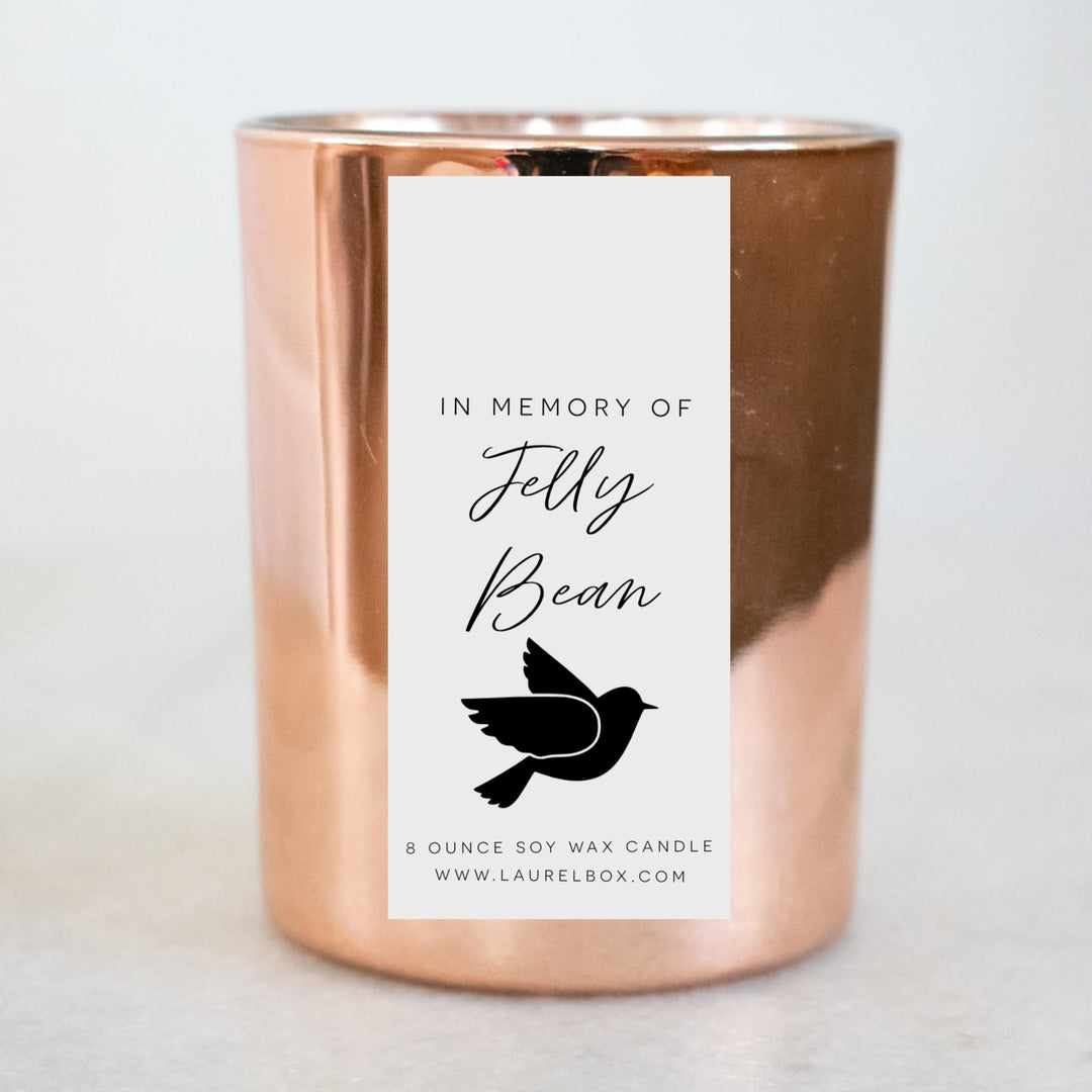A gold-coated candle with a white sticker, the text in Memory of Jelly Bean, and a bird symbol. Sitting on a marble surface