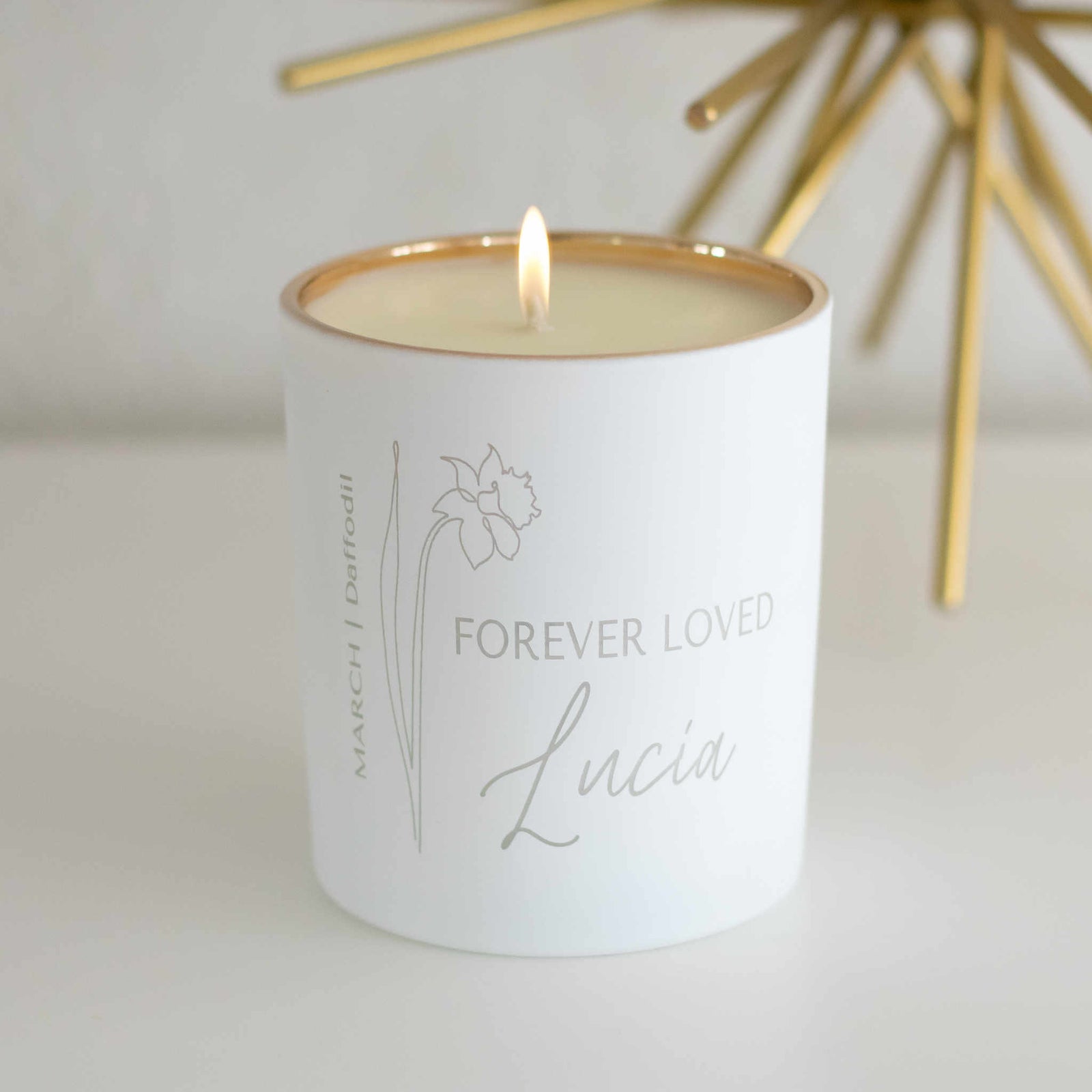 A white-coated candle with engraved text Forever Loved Lucia and a flower on the left. Sitting on a white surface.