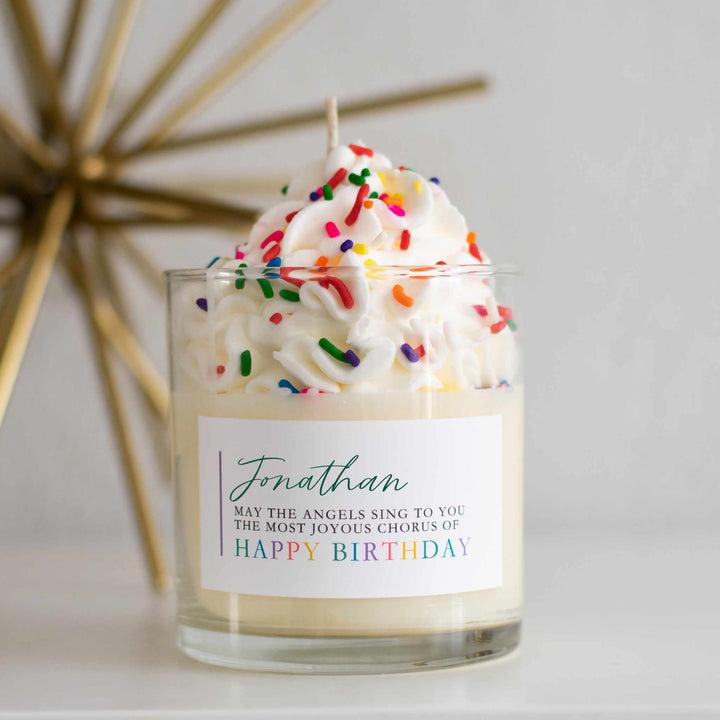 A glass candle labeled Jonathan May the angels sing to you the most joyous chorus of Happy Birthday with whipped cream and sparkles at the top. Sitting on a desk.