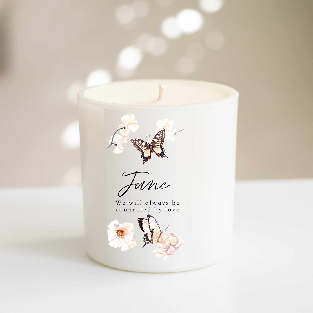 A white-coated candle with an engraved text of Jana, we will always be connected by love and butterflies and flowers scattered on it. Sitting on a white table