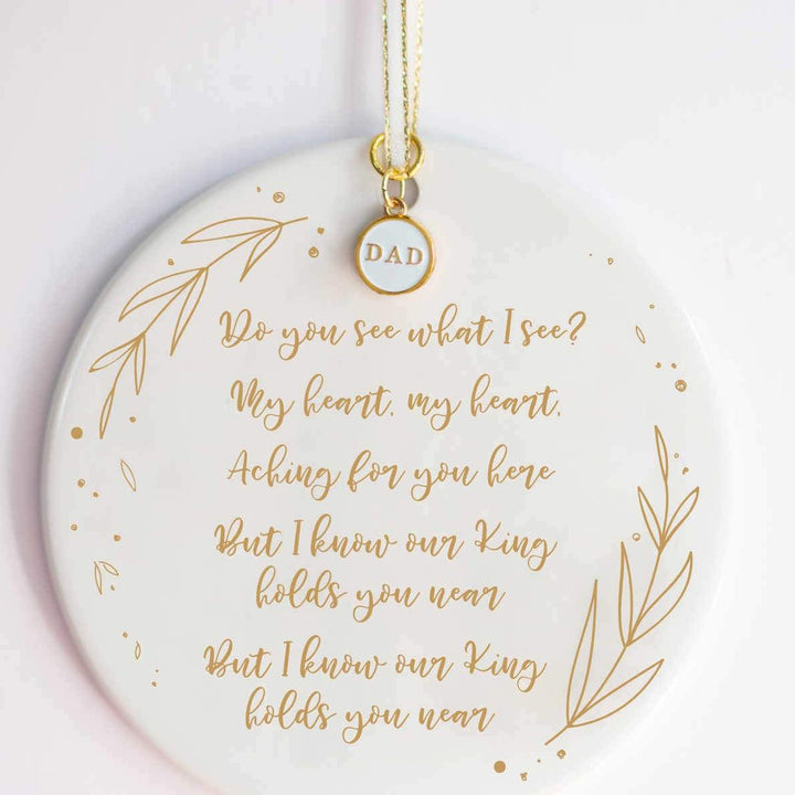 A white ceramic ornament with a "Do You See What I See" carol engraved with gold detailing and a white and gold enamel "DAD" charm at the top against a white background.