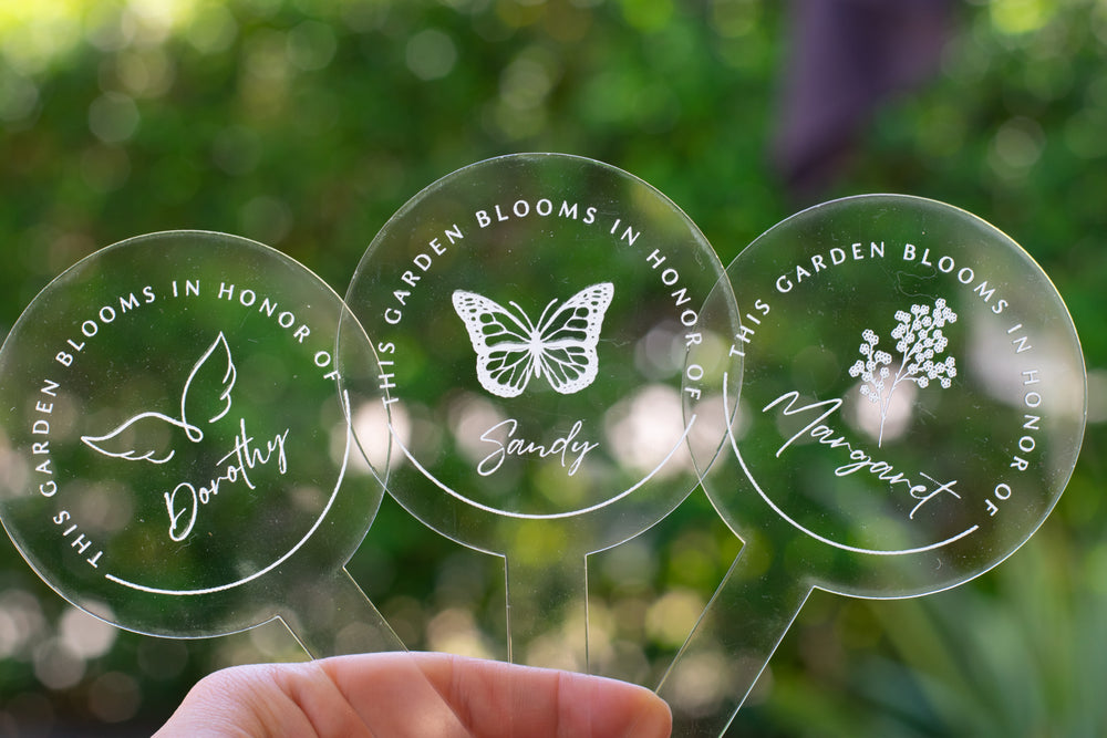 photo of three clear circular garden stakes - one that displays a flower and  "Margaret" at the center and the words "This Garden Blooms In Honor Of" around the flower, one that displays a butterfly and  "Sandy" at the center and the words "This Garden Blooms In Honor Of" around the butterfly and one that displays angel wings and  "Dorothy" at the center and the words "This Garden Blooms In Honor Of" around the angel wings