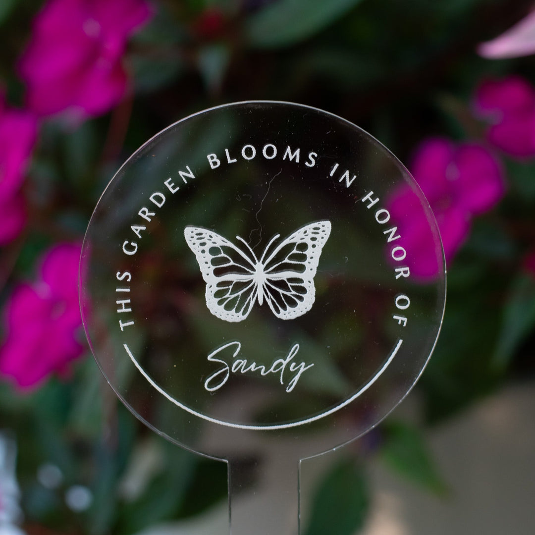 photo of a clear circular garden stake that displays a butterfly and  "Sandy" at the center and the words "This Garden Blooms In Honor Of" around the butterfly