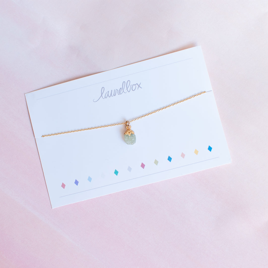 photo of a white piece of paper with laurelbox at the top with a gold chain necklace with a light green raw cut stone hanging at the bottom