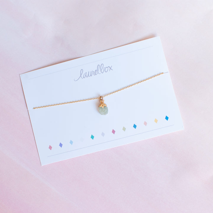 photo of a white piece of paper with laurelbox at the top with a gold chain necklace with a light green raw cut stone hanging at the bottom