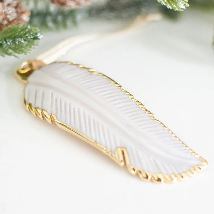 A close-up of a white mother-of-pearl feather ornament with gold edges and details. The ornament is laid flat on a white surface.
