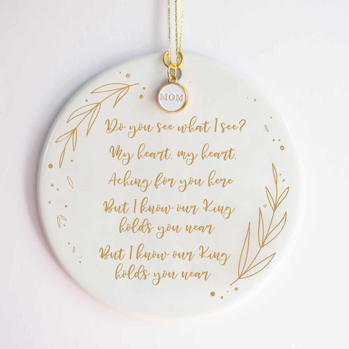 A white ceramic ornament with a "Do You See What I See" carol engraved with gold detailing and a white and gold enamel "MOM" charm at the top against a white background.