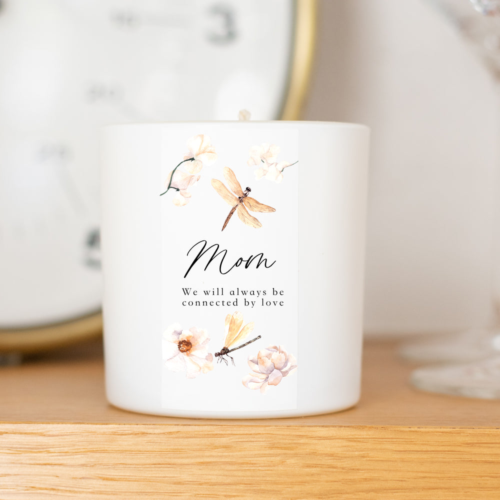 A white-coated candle with an engraved text of Mom, we will always be connected by love and butterflies and flowers scattered on it. Sitting on a wooden table with a clock in the background