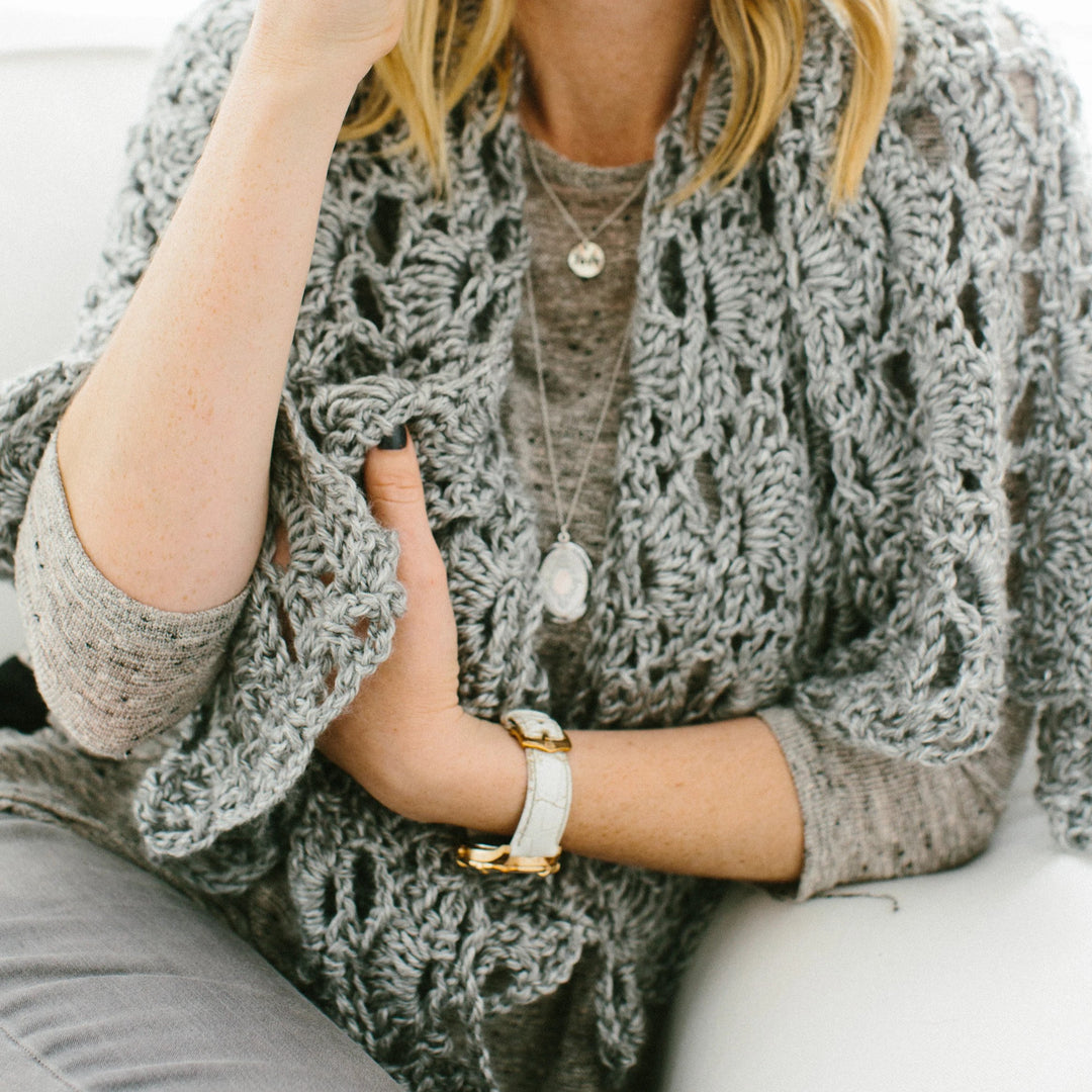 A close-up of a woman with blonde hair wearing a gray shirt, jeans, and a prayer shawl sitting on a white couch with her hand on her cheek.