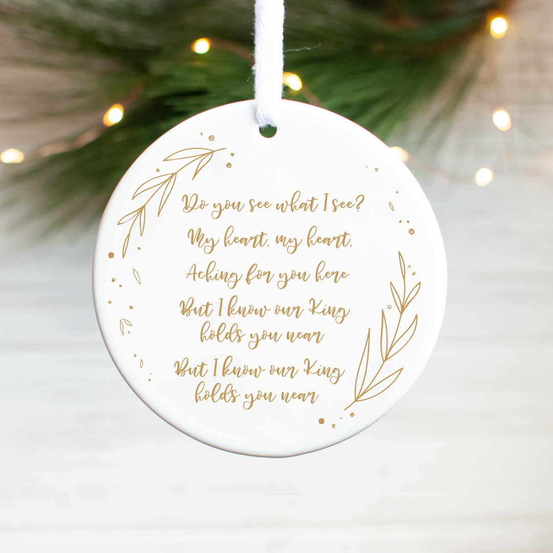 A white ceramic ornament with a "Do You See What I See" carol engraved in gold with gold detailing and pine branches and lights in the background.