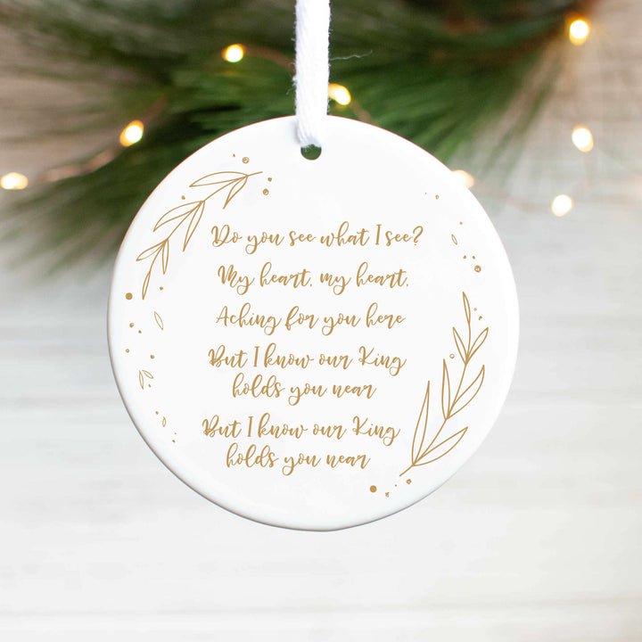 A white ceramic ornament with a "Do You See What I See" carol engraved in gold with gold detailing and pine branches and lights in the background.