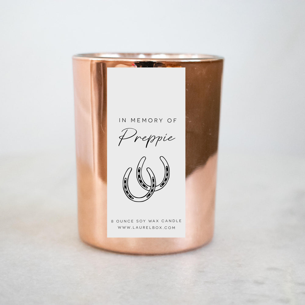 A gold-coated candle with a white sticker, the text in Memory of Preppie, and a horseshoe symbol. Sitting on a marble surface