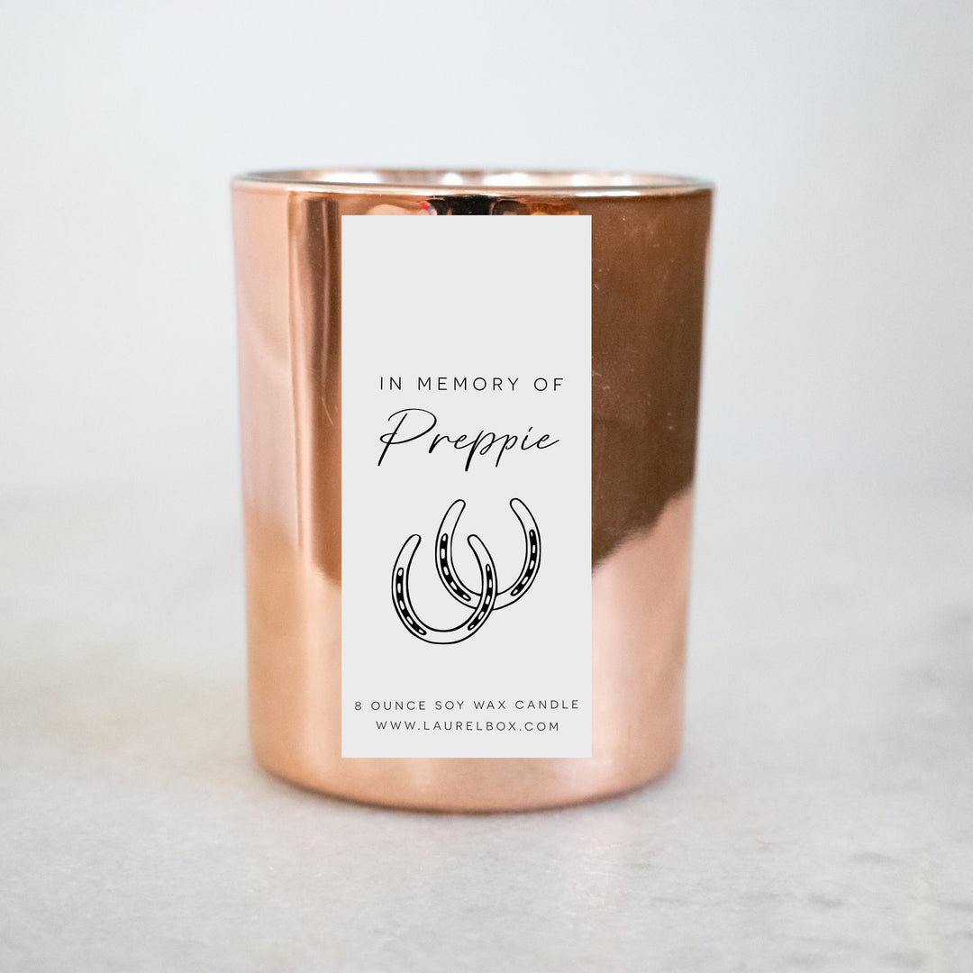 A gold-coated candle with a white sticker, the text in Memory of Preppie, and a horseshoe symbol. Sitting on a marble surface