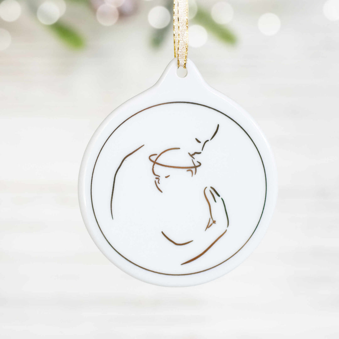 A close-up of a white ceramic ornament with an adult holding a baby with a halo design in metallic gold foil with pine branches in the background.