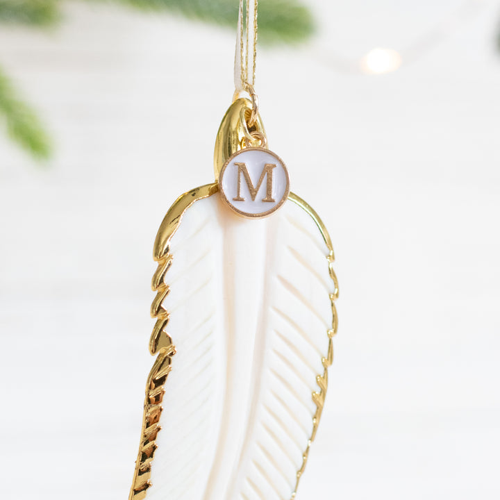 A close-up of a white mother-of-pearl feather ornament with gold edges and details and an "M" initial white and gold enamel charm at the top of the ornament.