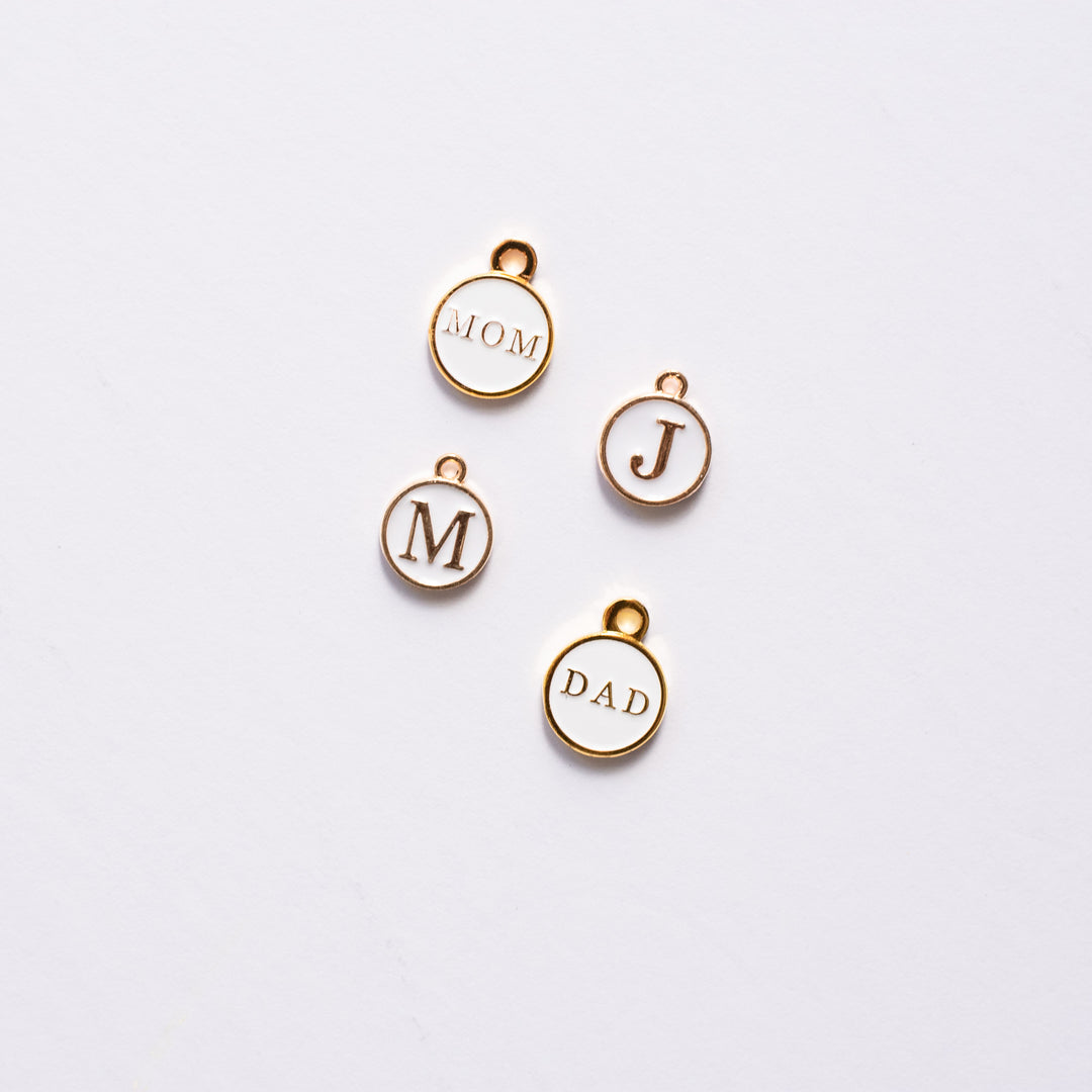 Four white and gold enamel initial charms are scattered against a white background.
