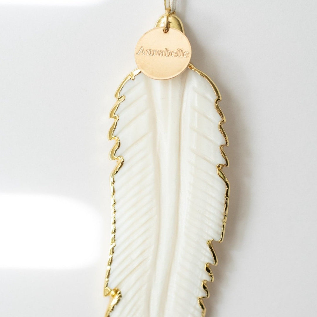 A close-up of a white mother-of-pearl feather ornament with gold edges and details and an "Annabelle" white and gold enamel charm at the top of the ornament. Highlighting the details and charm.