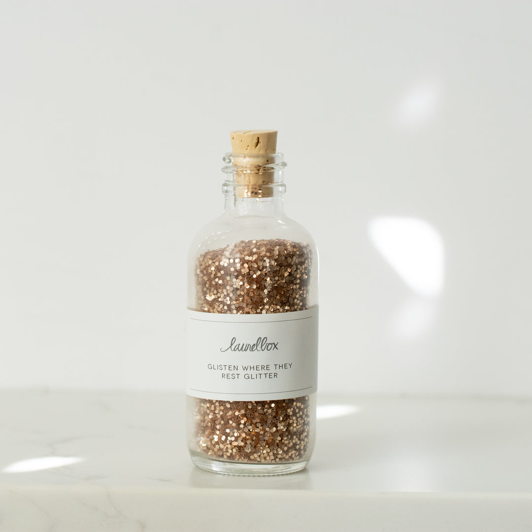 A bottle filled with gold glitter with a cork and white sticker with the text Glisten Where They Rest Glitter