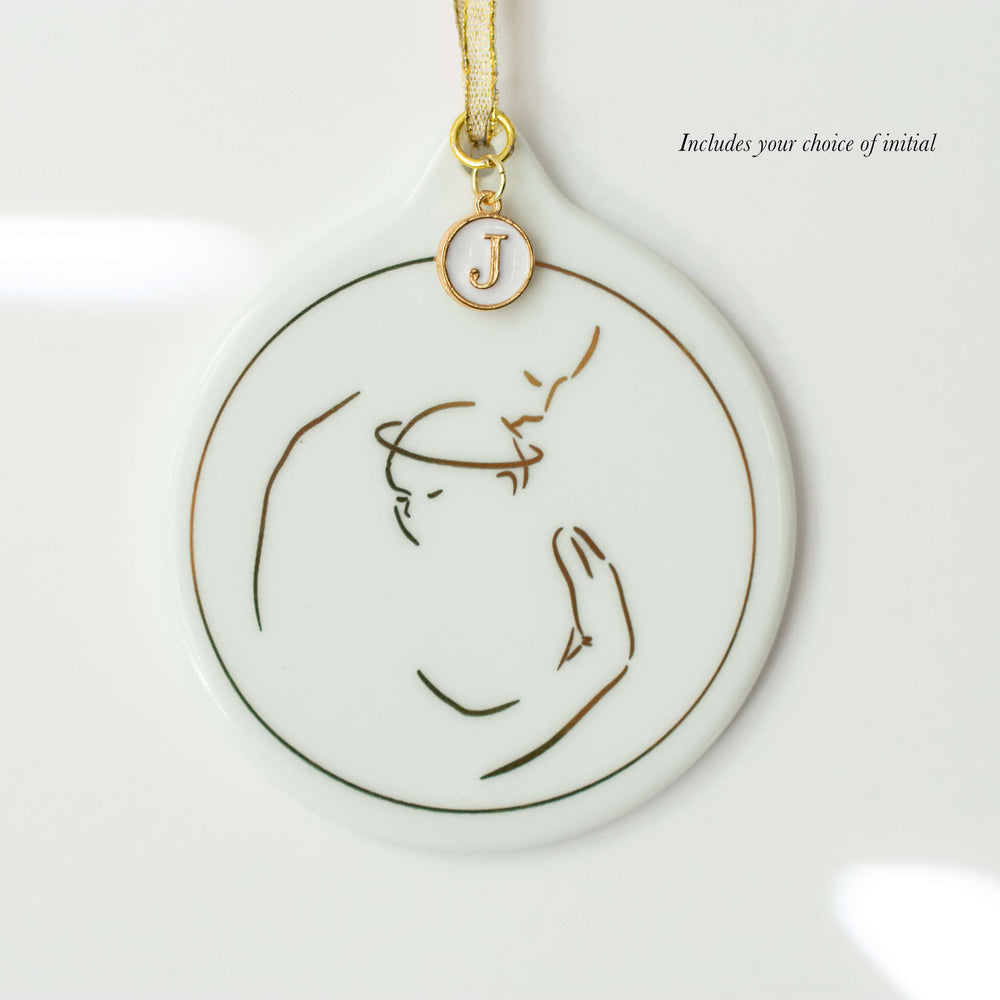 A white ceramic ornament with an adult holding a baby with a halo design in metallic gold foil with a white and enamel "J" initial charm against a white background. "Includes your choice of initial" text at the top right corner.