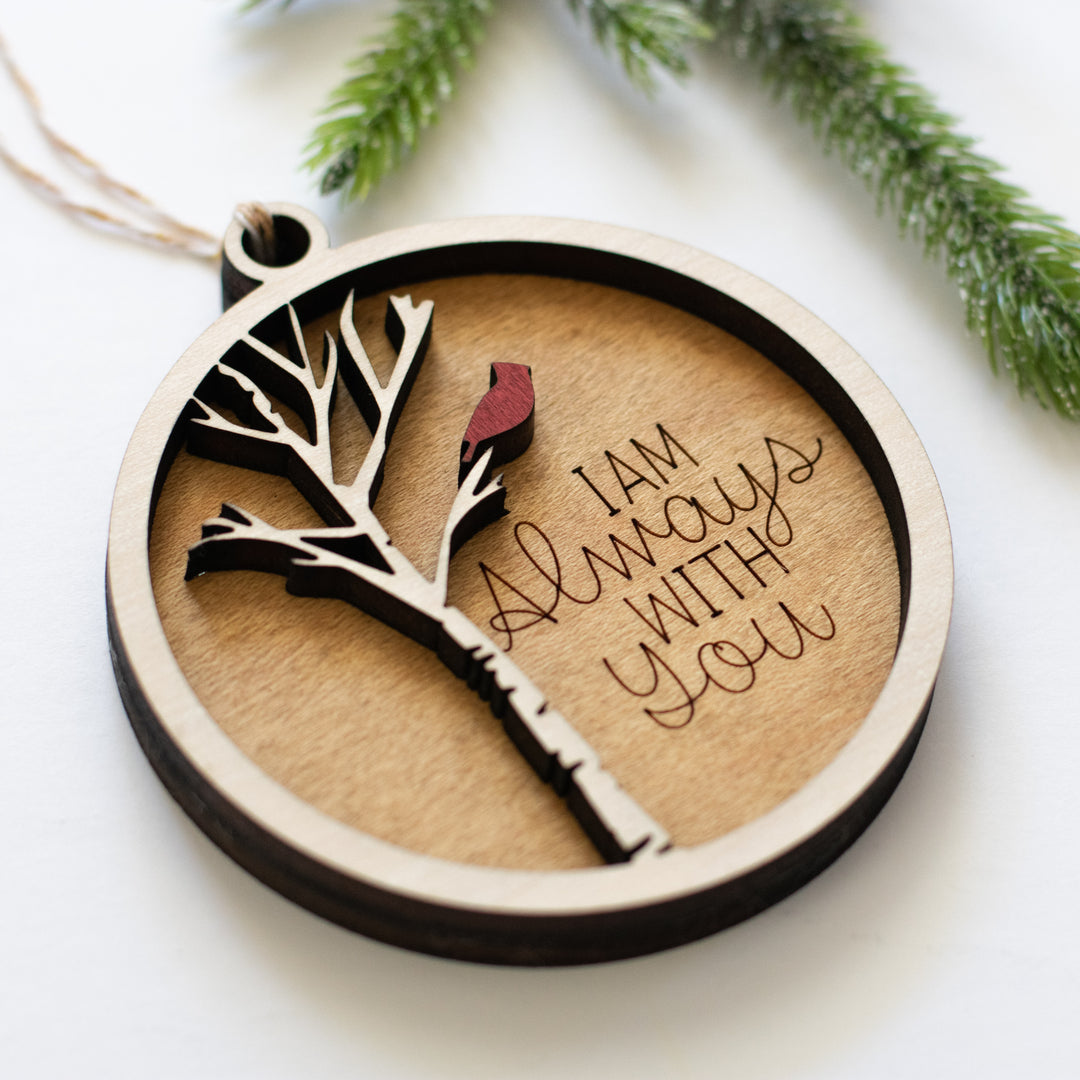 A close-up of a maple wood ornament featuring a cardinal on a white tree, with the text "I am always with you" engraved, resting on a white surface.