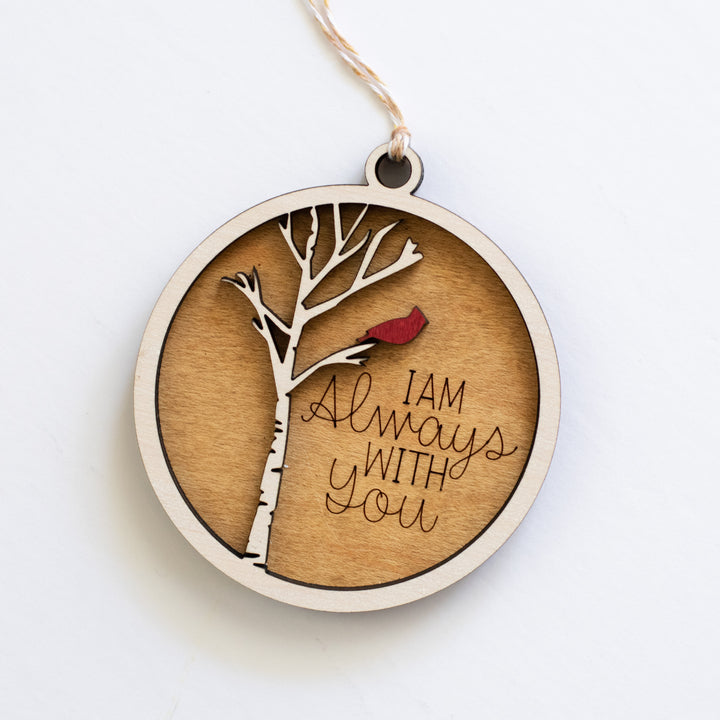 A close-up of a wooden ornament with a tree, cardinal, and I Am Always With You text engraved on it resting in the middle of a white surface.