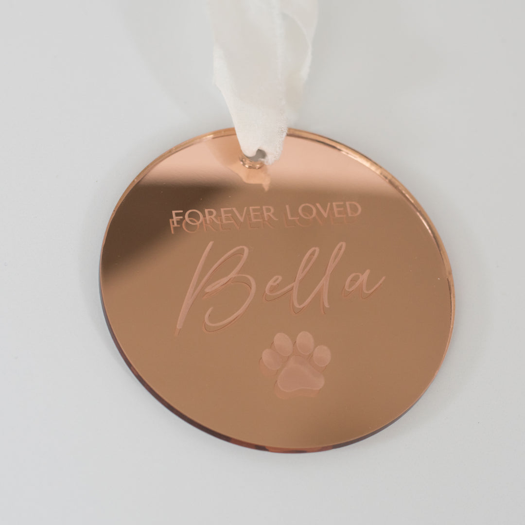A personalized gold mirrored ornament featuring the words "Forever Loved" and "Bella" with a paw print symbol engraved and a white ribbon at the top is set against a white background.