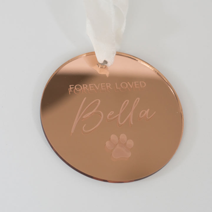 A personalized gold mirrored ornament featuring the words "Forever Loved" and "Bella" with a paw print symbol engraved and a white ribbon at the top is set against a white background.