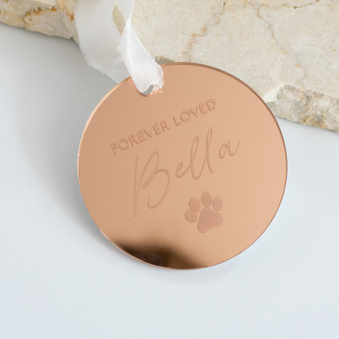 A personalized gold mirrored ornament featuring the words "Forever Loved" and "Bella" with a paw print symbol engraved and a white ribbon at the top is set against a marble surface on a white background.