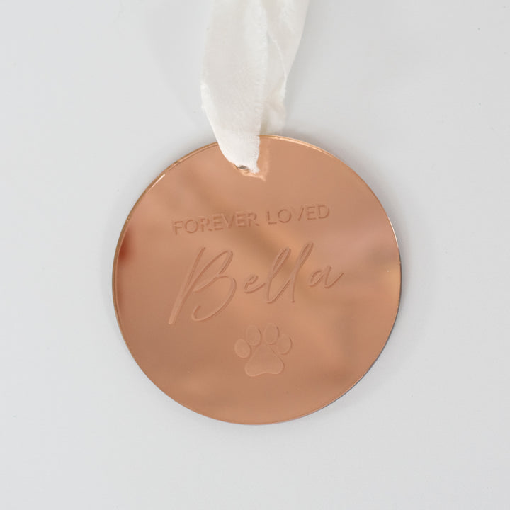A close-up of an angled personalized gold mirrored ornament featuring the words "Forever Loved" and "Bella" with a paw print symbol engraved and a white ribbon at the top is set against a white background.