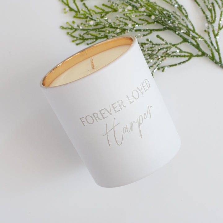A white-coated candle with engraved text Forever Loved Harper in gold. Tilted to the left against a white background