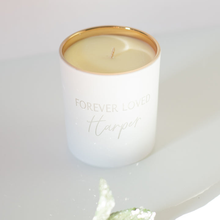 A white-coated candle with engraved text Forever Loved Harper in gold. Sitting against a white surface