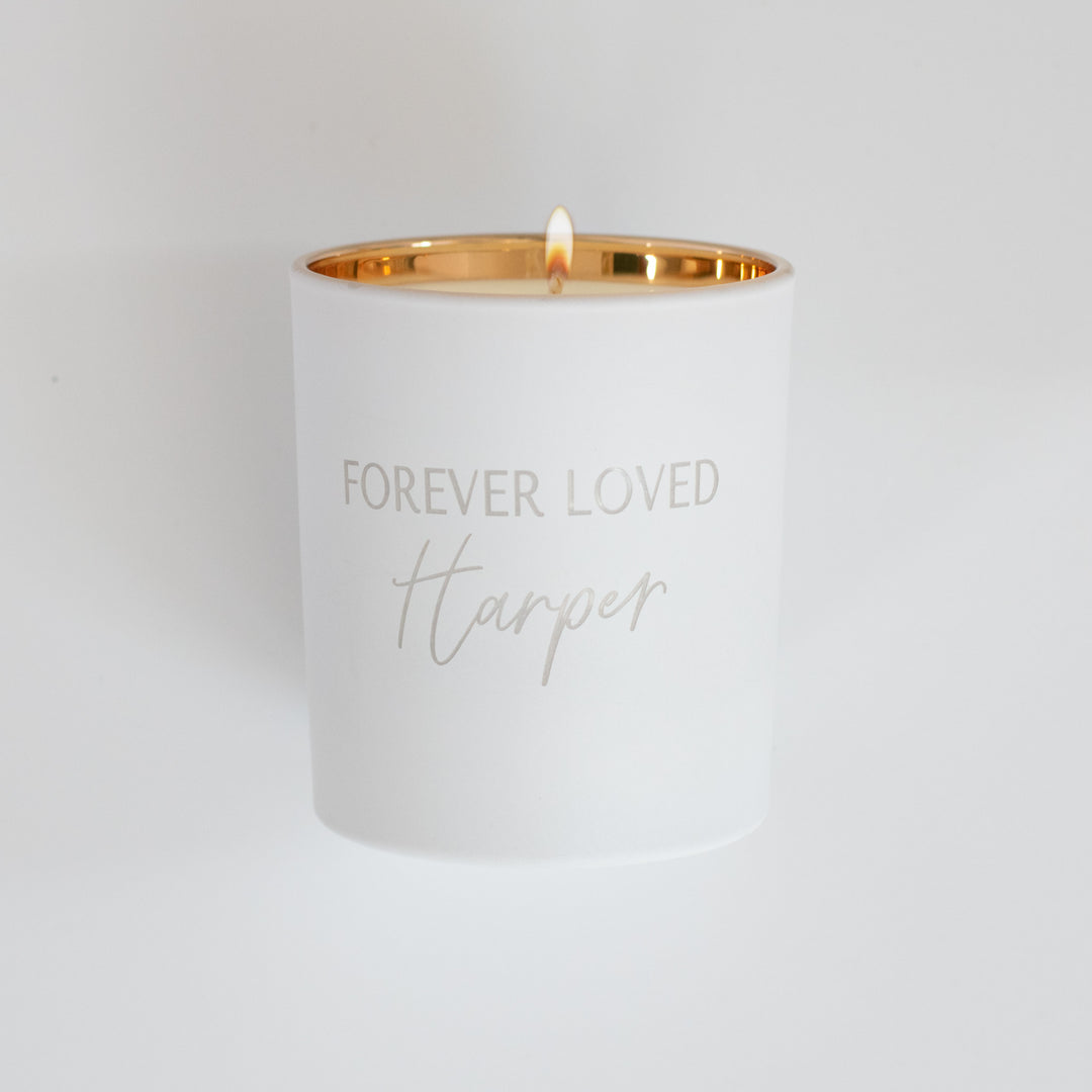 A white-coated candle with engraved text Forever Loved Harper in gold. Sitting against a white background