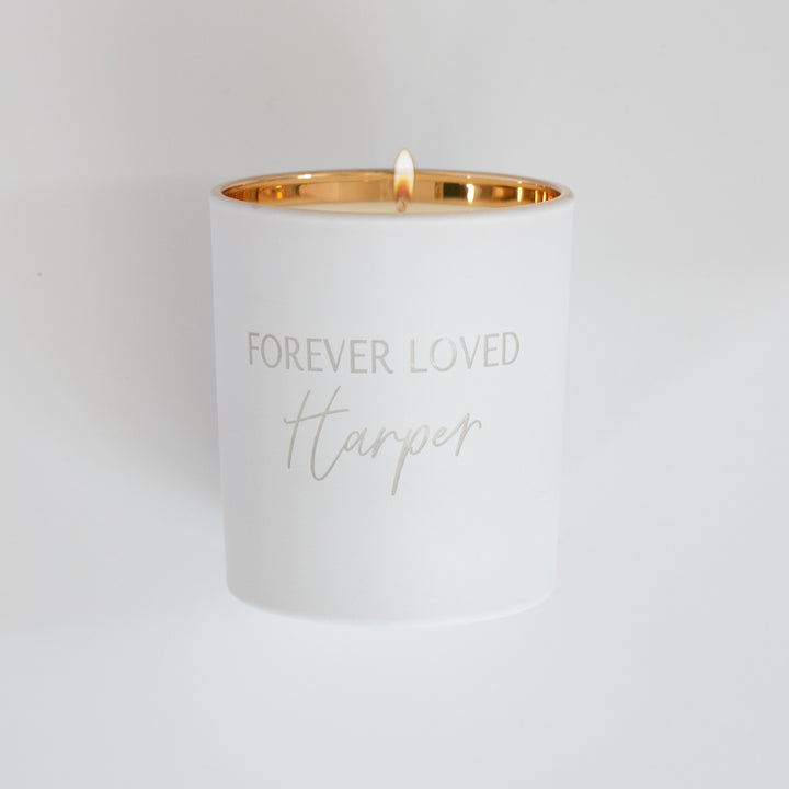 A white-coated candle with engraved text Forever Loved Harper in gold. Sitting against a white background
