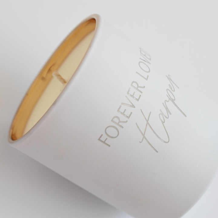 A close-up of a white-coated candle with engraved text Forever Loved Harper in gold. Angled against a white background
