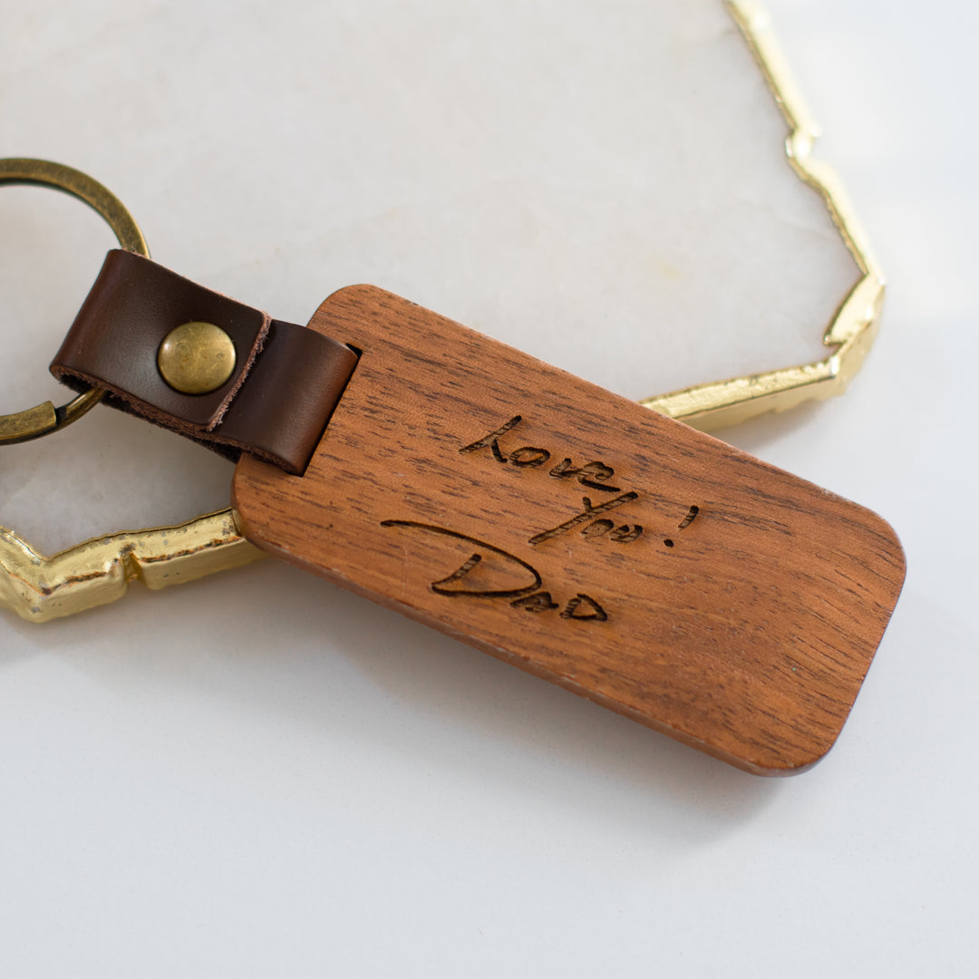 A close-up of a wooden keychain with Love You! Dad engraved on it, resting on a gold-framed granite surface.